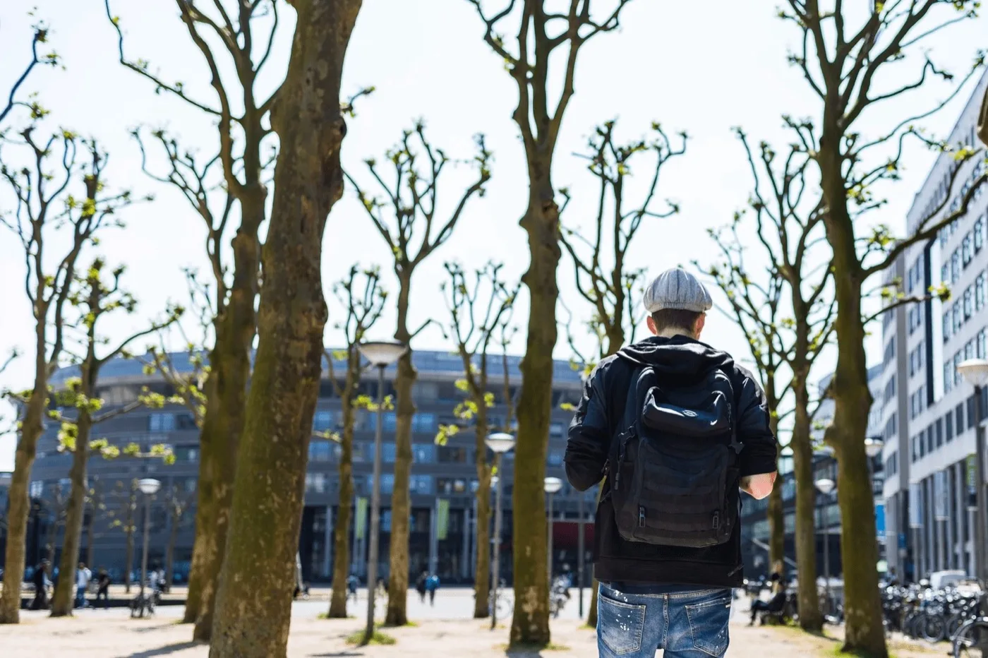 Man met rugzak loopt van de camera weg over een plein tussen kale bomen